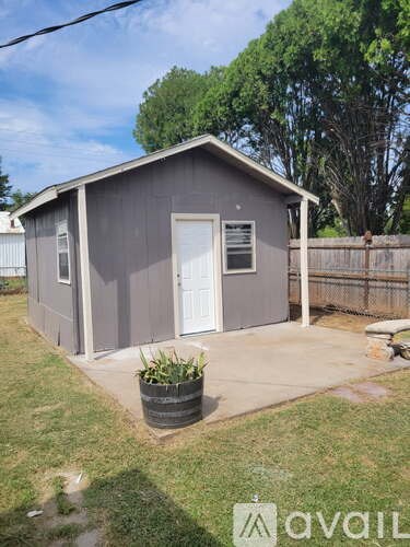 A gray shed with a white door and a window is surrounded by a wooden fence and has a potted plant in front.