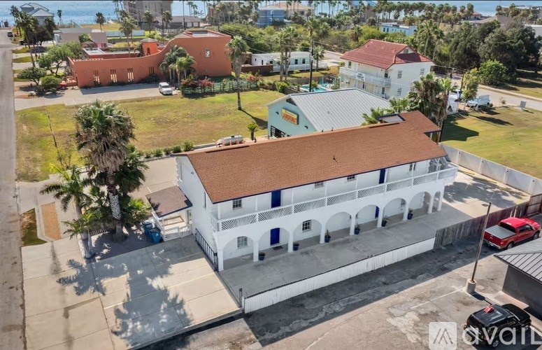 A white building with a red roof is surrounded by other buildings and palm trees.