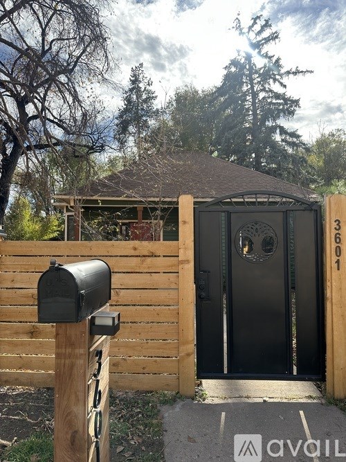 A black mailbox is on a wooden post in front of a wooden fence and a black gate.