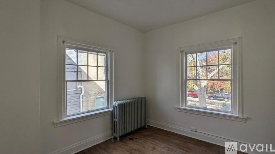 A room with two windows, one facing a building and the other a tree-lined street.