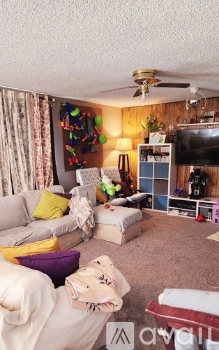 A living room with a grey couch, a white chair, a white ottoman, a white blanket, a white pillow, a white wall, a white ceiling, a white ceiling fan, a white door, a white cur