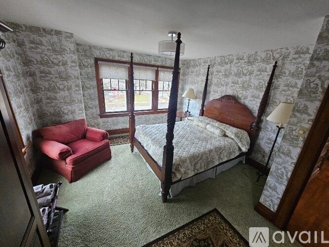 A bedroom with a four poster bed, a red chair and a window.