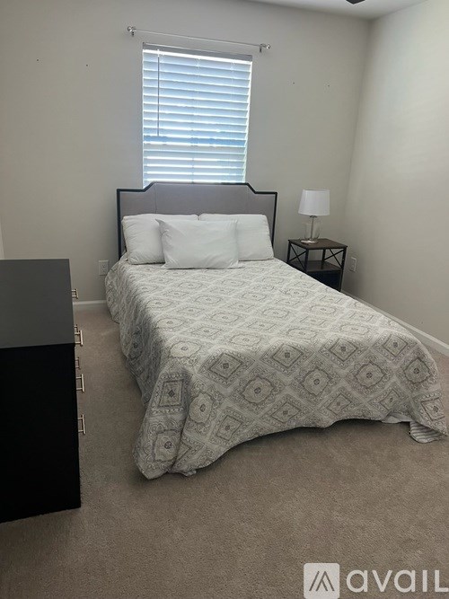 A bed with a white comforter and a black headboard is in a room with a window and a nightstand with a lamp.