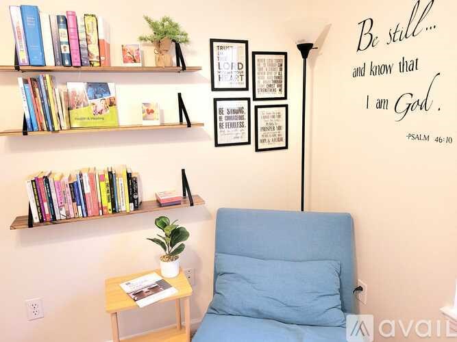 A room with a blue chair, a shelf with books, a small table with a plant, and a wall with a quote and pictures.