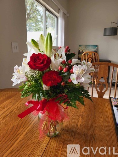 A bouquet of red roses, white lilies, and other flowers in a clear vase with a red ribbon sits on a wooden table.