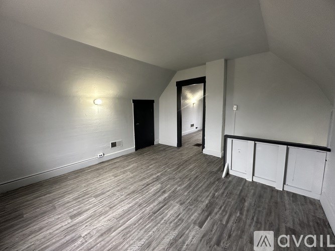 A room with wooden flooring and white walls, with a door on the right and a light switch on the left wall.