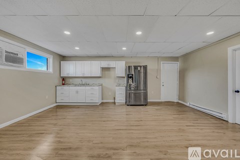 A spacious room with wooden flooring and white walls, featuring a kitchenette with white cabinets and a stainless steel refrigerator.