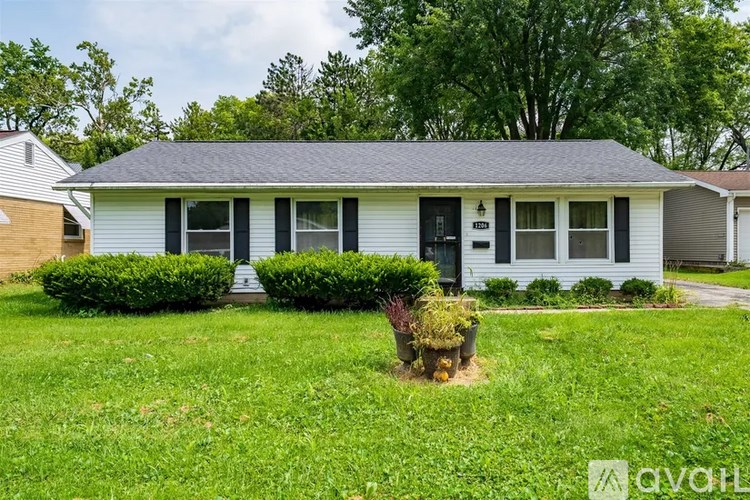 A white house with a black door and windows is surrounded by greenery.