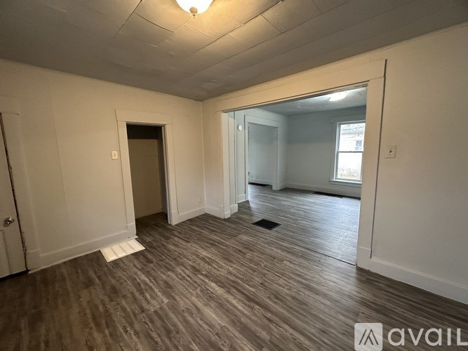 A room with wooden flooring and white walls, with a door on the left and a window on the right.