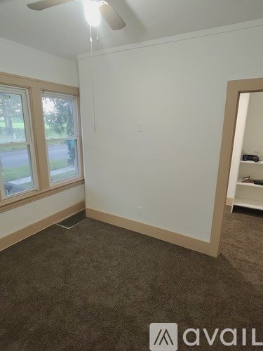 A room with a carpeted floor, a ceiling fan, and a window overlooking a street.