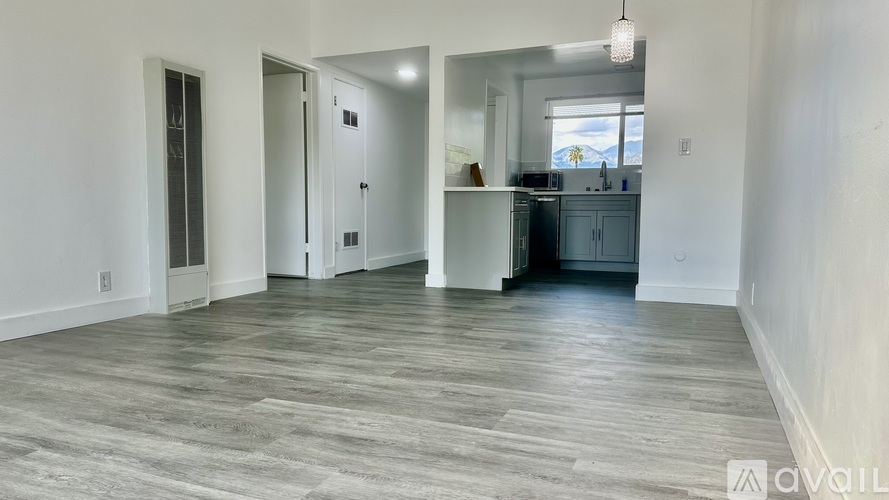A room with wooden flooring and white walls, with a kitchen visible in the background.