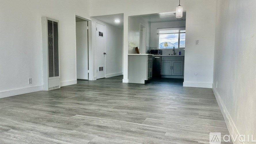A room with wooden flooring and white walls, with a kitchen visible in the background.