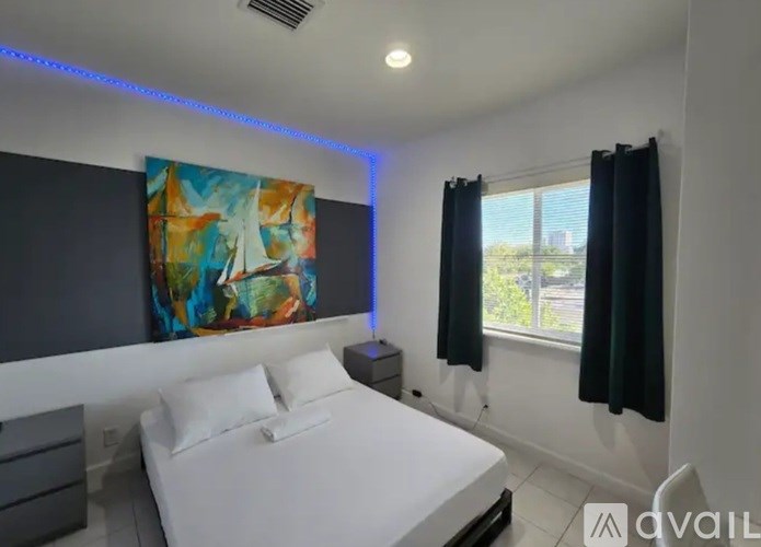 A bedroom with a large bed, a painting on the wall, and a window with black curtains.