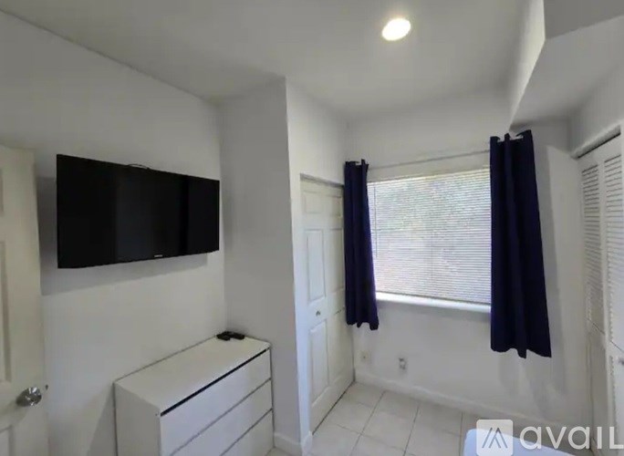 A room with a black television, a white dresser, and a window with black curtains.