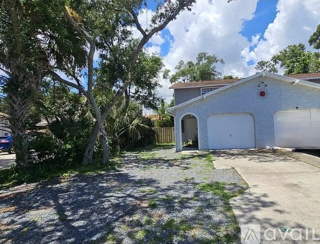 A white garage with a red decoration on the roof is surrounded by trees and a gravel driveway.