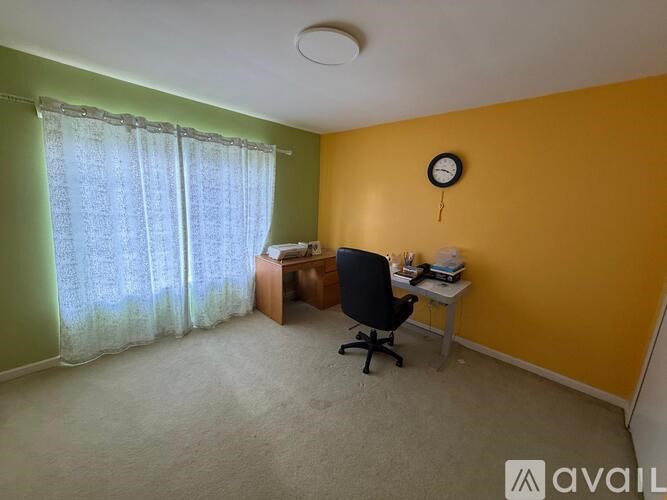 A room with a desk and chair, a window with curtains, and a clock on the wall.