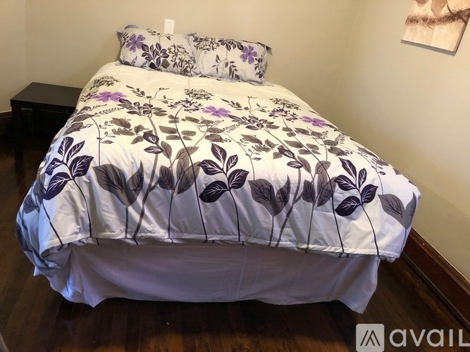 A bed with a white and purple comforter and a small black table beside it.