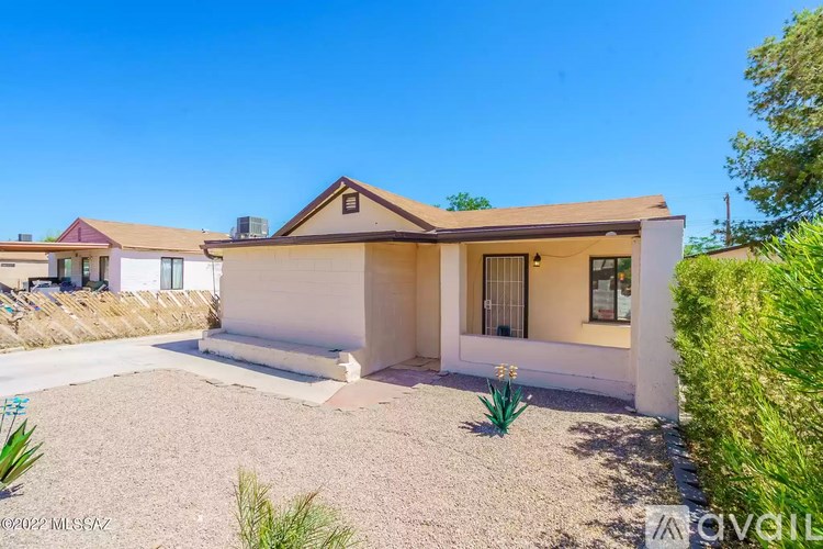 A house with a brown roof and white walls is surrounded by a gravel driveway and some greenery.