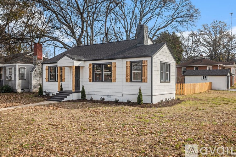 A white house with a black roof and a chimney is surrounded by trees and other houses.