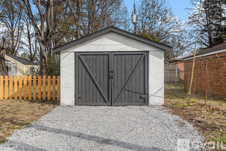 A small white building with a black door is surrounded by a gravel driveway and a wooden fence.