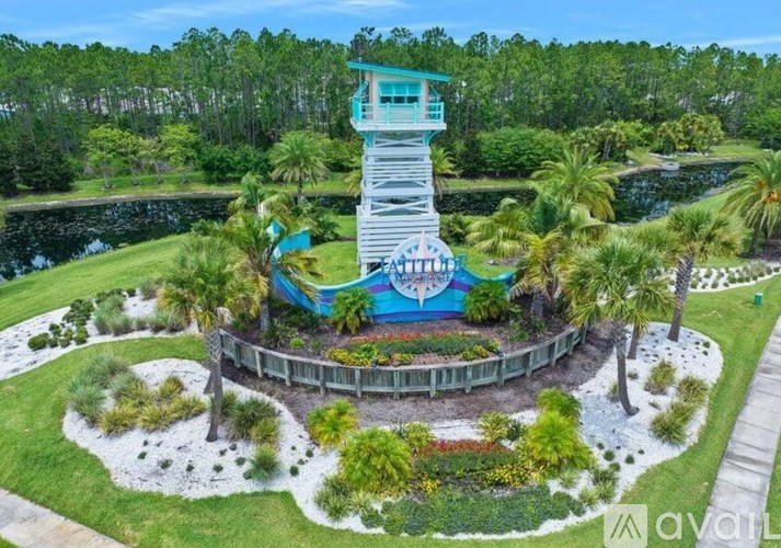A blue and white lifeguard tower sits atop a circular platform with a white fence and a variety of plants and trees surrounding it.