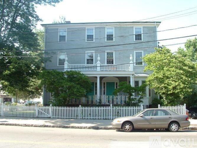 A silver car is parked in front of a grey house with a white picket fence.