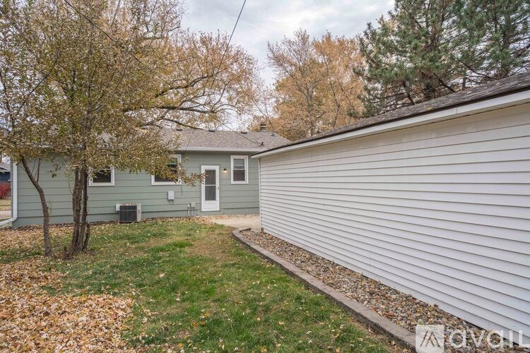 A house with a grey siding and a white door is surrounded by a grassy area with a tree and fallen leaves.