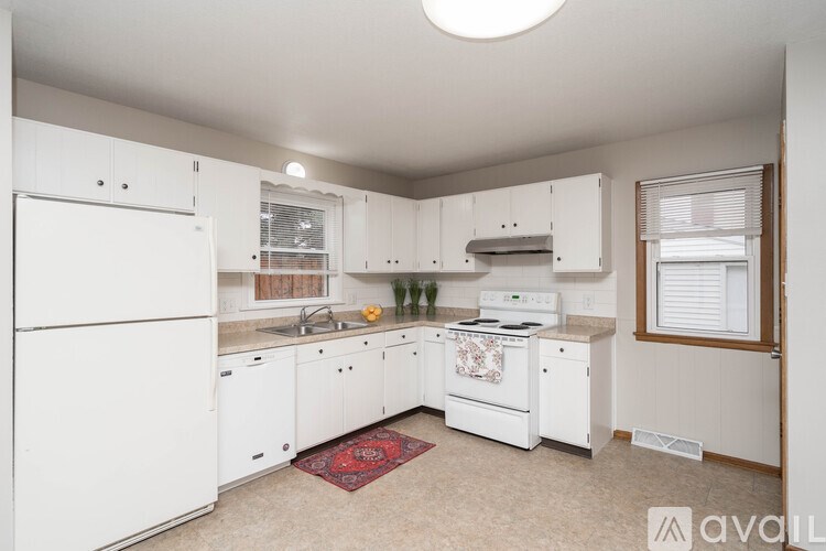 A kitchen with white appliances and cabinets, a rug on the floor, and a window with blinds.