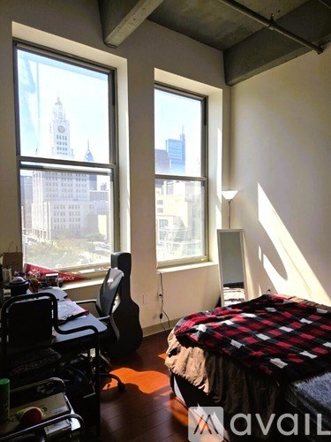 A room with a bed, desk, and chair with a window overlooking a city.