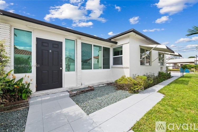 A white house with a black door and windows is surrounded by greenery and a gravel path.