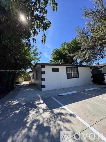 A white house with a black door and windows is surrounded by trees and has a driveway in front.