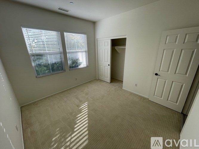 A room with a carpeted floor, two windows with blinds, and a door leading to another room.