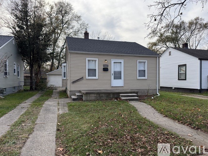 A small house with a grey roof and a white door is surrounded by a grassy area and a concrete pathway.