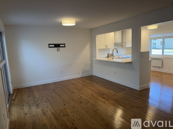 A room with wooden floors and white walls, with a kitchen area visible in the background.