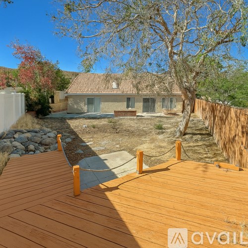 A wooden deck leads to a house with a stone wall and a tree.