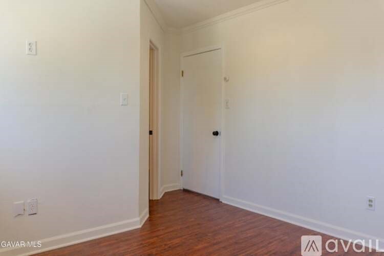 A room with white walls and wood flooring, with a door on the right and a light switch on the left wall.