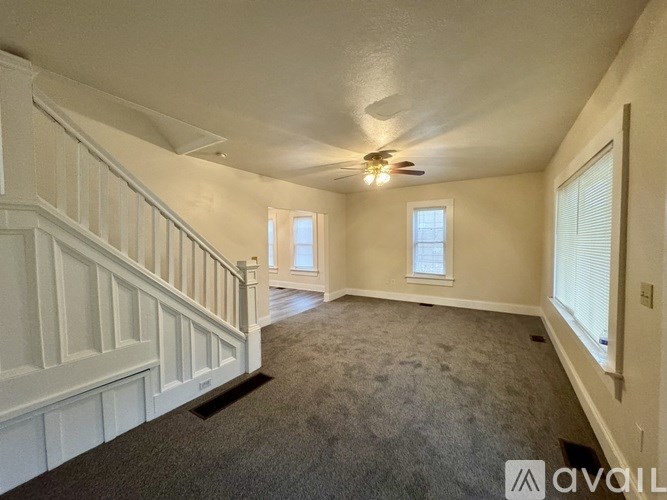 A spacious room with a carpeted floor, a staircase with white railings, and a ceiling fan.