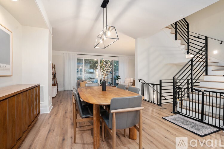 A dining room with a wooden table and chairs, a staircase with a metal railing, and a window with a view of trees.