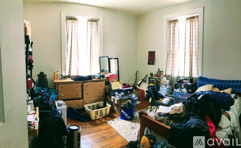A cluttered bedroom with a bed, desk, and clothing scattered about.
