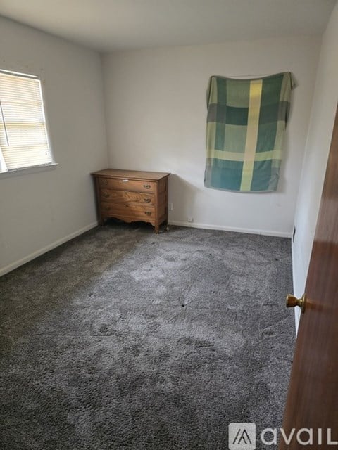 A room with a carpeted floor, a wooden chest of drawers, and a window with blinds.