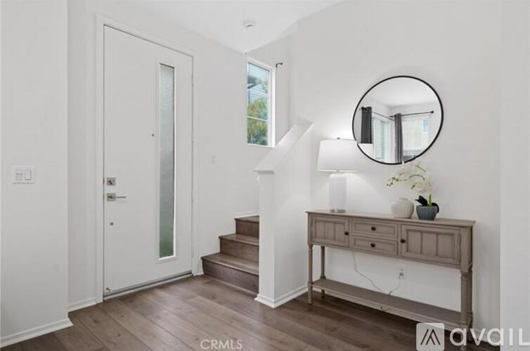 A hallway with a wooden floor, a white door, a staircase, a table with a lamp and a mirror.