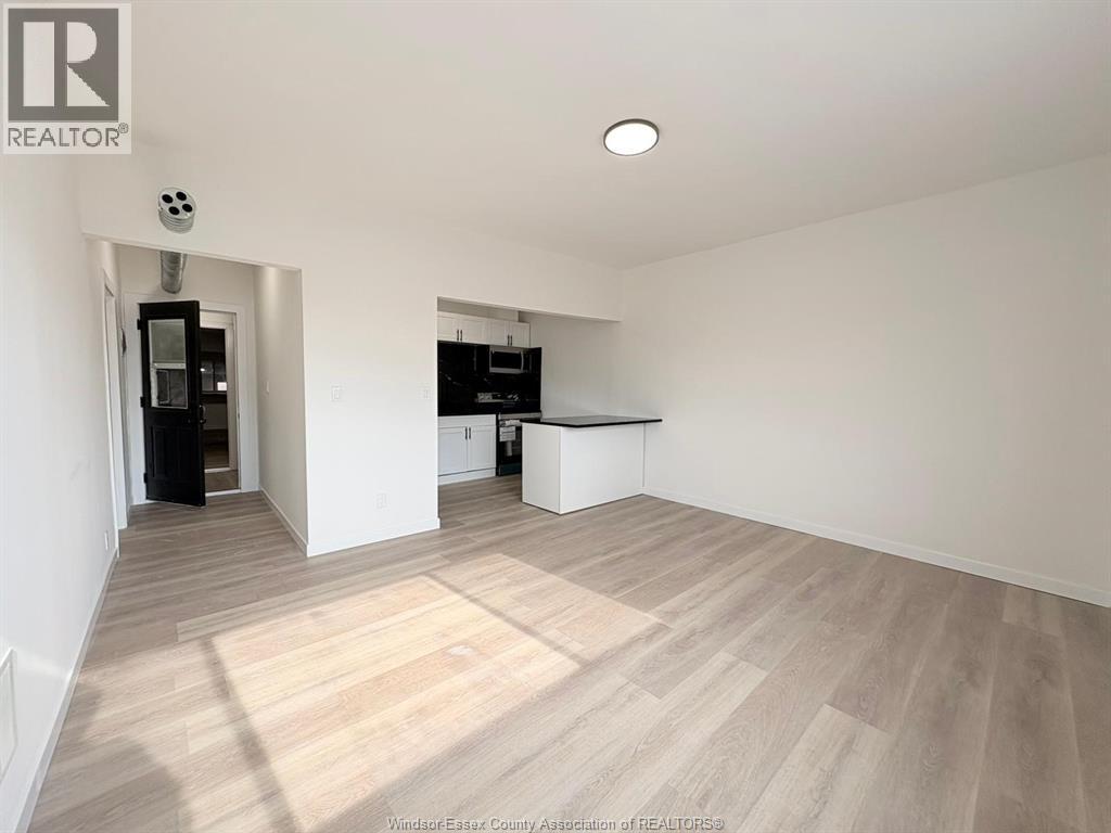 A spacious room with wooden flooring and white walls, with a kitchen area visible in the background.
