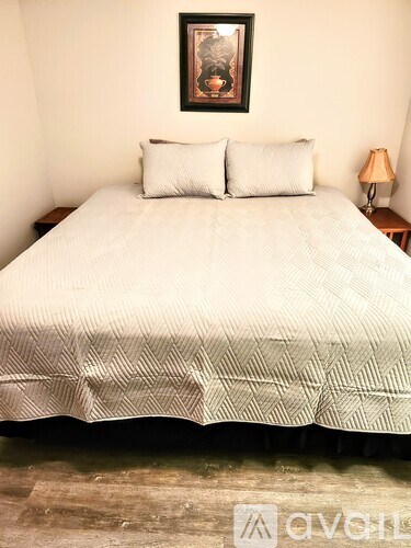 A bed with a white comforter and two pillows sits in a room with a framed picture on the wall.