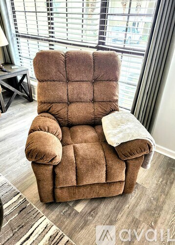A brown recliner chair with a white pillow is in front of a window with blinds.