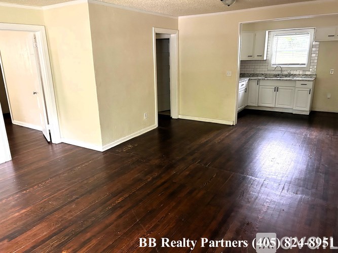 A room with wooden floors and white walls, with a kitchen area visible in the background.