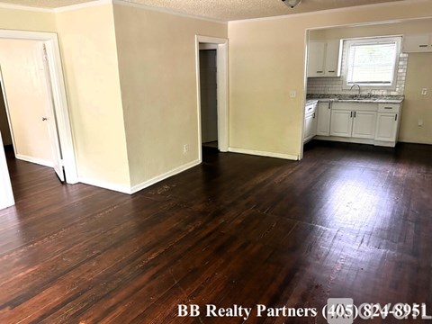 A room with wooden floors and white walls, with a kitchen area visible in the background.