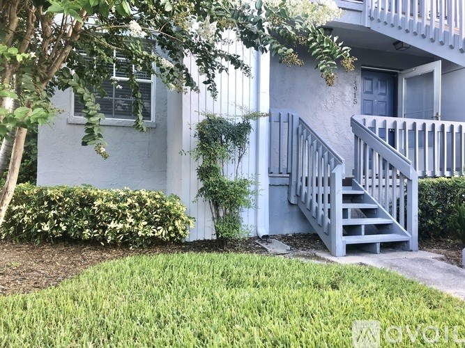 A white house with a grey staircase and a tree in front.