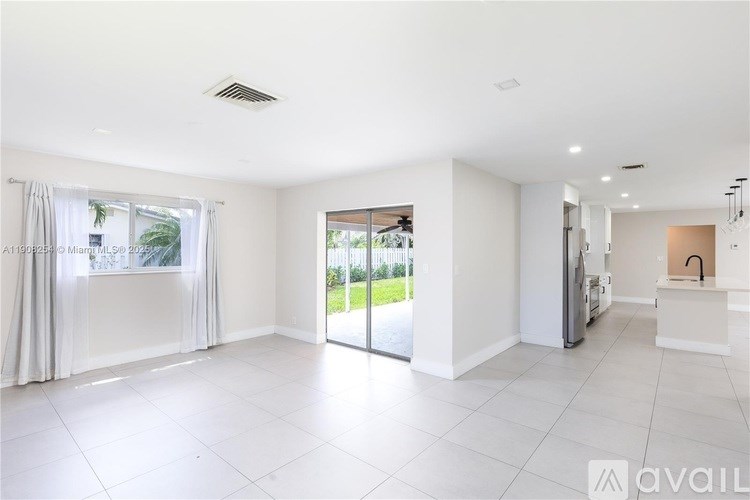 A spacious, empty room with white walls and flooring, a sliding glass door, and a kitchen area with a sink and stove.