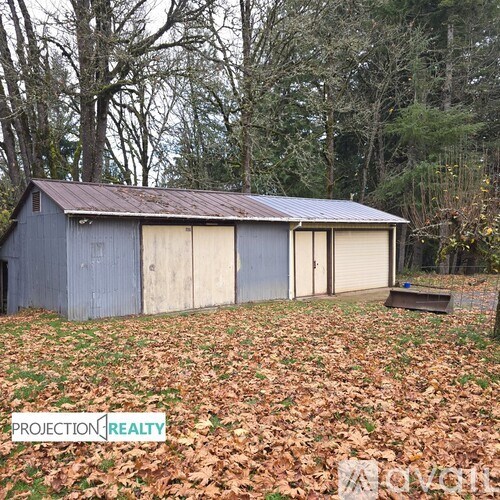 A blue barn with a metal roof is surrounded by trees and fallen leaves.