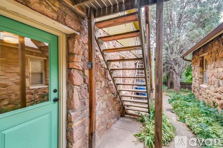 A green door is on the left of a stone wall with a wooden staircase leading up to a porch.